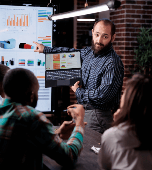 startup-entrepreneur-holding-laptop-with-sales-presentation-charts-pointing-wall-screen-tv-late-night-meeting-caucasian-man-presenting-marketing-strategy-coworkers-working-overtime 1 (1)
