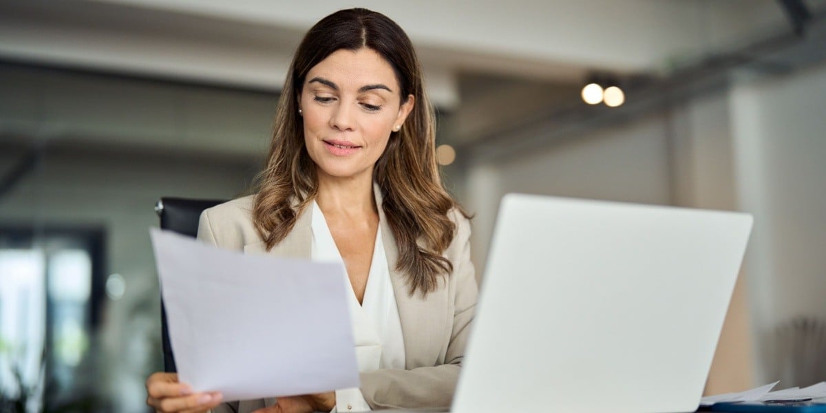 Businesswoman reading a sheet of paper sitting in front of her laptop